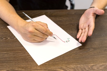 Businessman sitting at table and holds pen in right hand. There are sheet of paper with a trading...