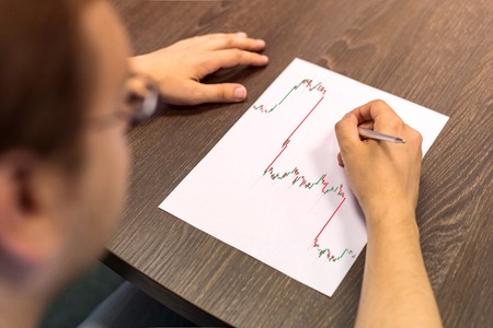 Businessman sitting at table and holds pen in right hand. There are sheet of paper with a trading...