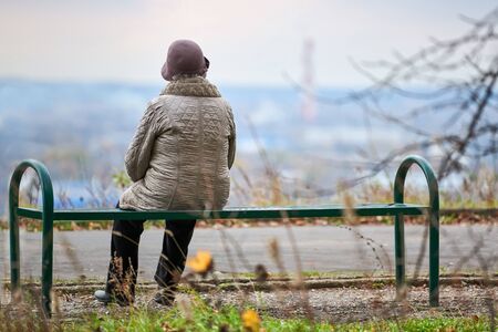Old woman sitting on bench in autumn city park. Lonely widow looking deep at city, self-reflection. Grandmother dreaming about her family concept.の写真素材