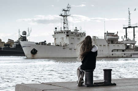 Young girl back view looking at ships. Woman dreaming about sea travel. Long-haired female sitting on moorings, ships in port background. Loneliness conceptの写真素材