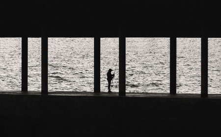 Silhouette of fisherman with fishing rod on pier. Lonely old fisherman on sea background. Black and white photo.の写真素材
