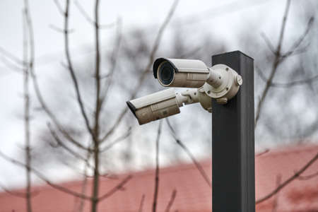 IP CCTV security camera on pole, gray sky, trees and roof background, cityscape. Protected area, private property protection.の写真素材