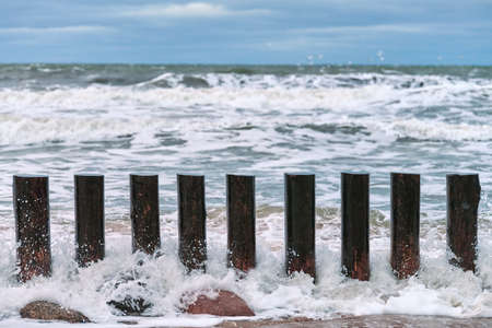 High wooden breakwaters in splashing sea waves, beautiful cloudy sky, close up view. Long poles or groynes in stormy sea.の写真素材