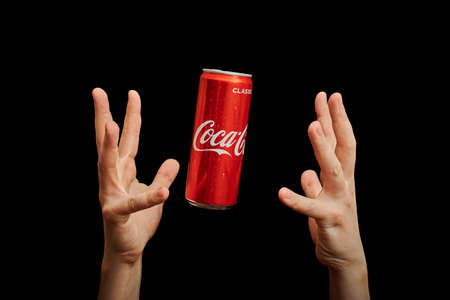 KALININGRAD, RUSSIA - MARCH 13, 2021 - Hands catch Coca Cola can, black background. Classic coke jar, carbonated soft drink. Manufactured by The Coca-Cola Company.のeditorial素材