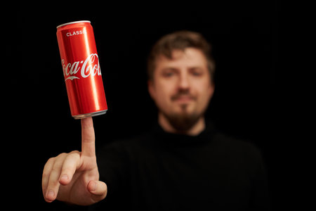 KALININGRAD, RUSSIA - MARCH 13, 2021 - Man with Coca Cola can on finger, black background. Classic coke jar, carbonated soft drink. Manufactured by The Coca-Cola Company.のeditorial素材