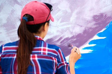 Female artist painting on wooden canvas board at outdoor festival, young adult woman painter creating abstract picture in purple, violet, blue and white colors moving paint brush, back viewの写真素材