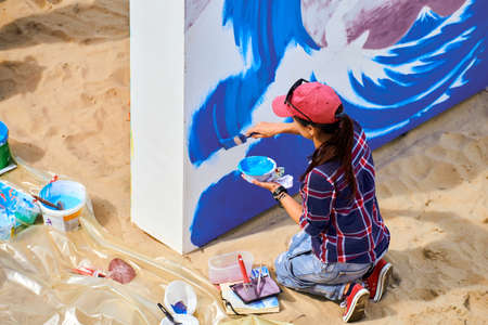 Female artist painting on wooden canvas board at outdoor festival, young adult woman painter creating abstract picture in purple, violet, blue and white colors moving paint brush, side viewの写真素材