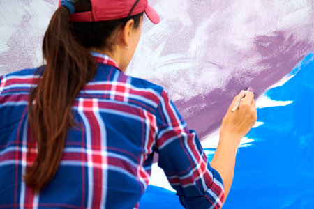 Female artist painting on wooden canvas board at outdoor festival, young adult woman painter creating abstract picture in purple, violet, blue and white colors moving paint brush, back viewの写真素材