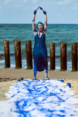 Young woman performance artist in dark blue dress smeared with blue gouache paints happily dancing...