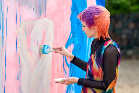 Svetlogorsk, Russia - 08.14.2021 - Woman artist with short multicolored hair in bright colorful clothes painting beautiful picture with paint brush. Oil painting on large canvas with blue, pink colorsのeditorial素材