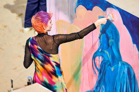 Svetlogorsk, Russia - 08.14.2021 - Woman artist with short multicolored hair in bright colorful clothes painting beautiful picture with paint brush. Oil painting on large canvas with blue, pink colorsのeditorial素材