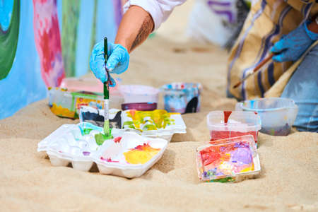 Svetlogorsk, Russia - 08.15.2021 - Woman painter in blue gloves choosing colors to paint picture with paint brush. Mixing paints of different colors. Acrylic painting, outdoor art performance festivalのeditorial素材