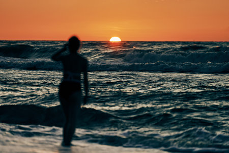 Female silhouette in blue sea waves at beautiful summer sunset, half sun below horizon. Adult girl into sea waves watching beautiful sunset, horizon landscape, summer beachfront holidayの写真素材