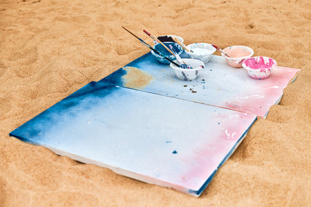 Two painted canvas in blue and pink colors with palettes of paints lying on sandy beach, artist's palette at outdoor art festival. White drawing canvas with paint in bowls, art performance conceptの写真素材
