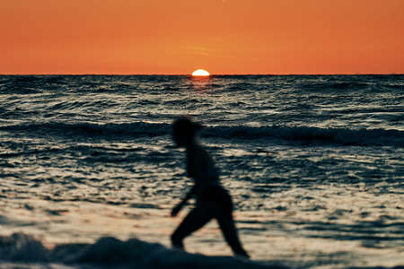 Female silhouette in blue sea waves at beautiful summer sunset, half sun below horizon. Young woman walking into sea waves watching beautiful sunset, setting sun on horizon, summer beachfront holidayの写真素材