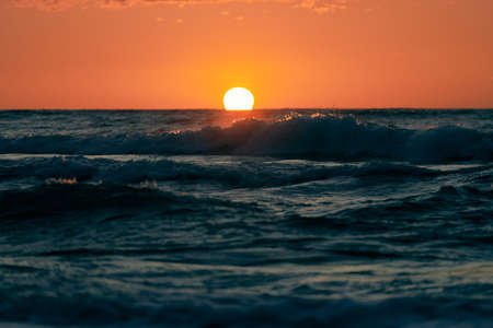 Half sun below horizon over blue sea waves, beautiful sunset over sea. Turquoise sea waves in dramatic sunshine from orange sun in sunset sky, breathtaking seascape with atmospheric sea wavesの写真素材