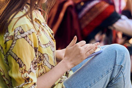 Sitting woman in yellow blouse and blue jeans using smartphone for messaging, outdoor resting. Young female holding smart phone for scrolling social media content and textingの写真素材