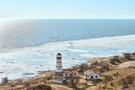 Atmospheric romantic view to white red lighthouse with farm utility houses in Merzhanovo, Rostov on Don Russian region, half frozen Azov sea horizon background. Old wooden lighthouse on Azov seashoreの写真素材