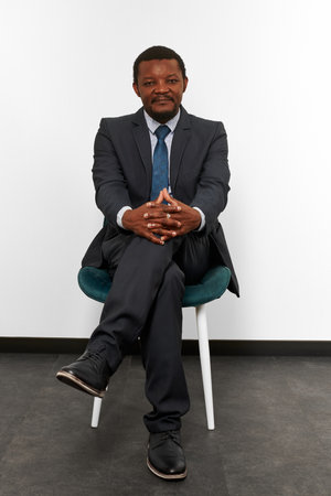 Smiling african american black man in business suit sitting on chair with crossed fingers, white wall background. Full size portrait of happy black man chief executive officerの写真素材