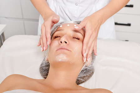 Cosmetologist rubbing moisturizing cream mask into woman face skin for rejuvenation in beauty salonの写真素材