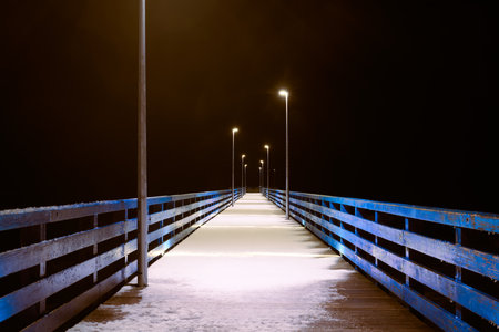 Sea pier covered with snow in night yellow lights from lampposts, mysterious path in darknessの写真素材