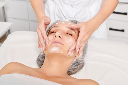 Beautician rubbing moisturizing cream mask into woman face skin for rejuvenation in beauty salonの写真素材