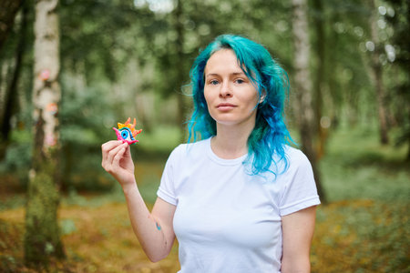 Young woman with turquoise dyed hair in white T shirt holds handmade colorful eye amulet in parkの写真素材