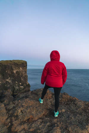 the back of a woman standing by a cliff near the ocean at sunsetの写真素材
