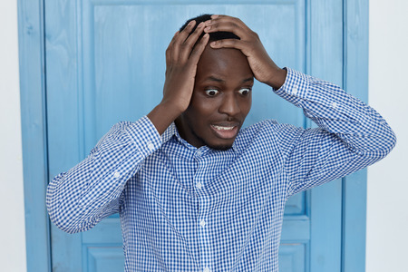 Funny wide-eyed young Afro-American man in checkered shirt having scared crazy look, raising brows. Remember important meeting that he missed due to bad memory or overworking.の写真素材