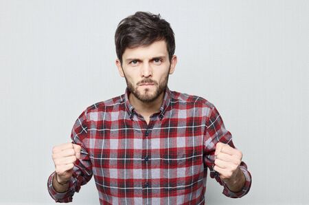 Studio close up of Caucasian male who holds his fists in anger and threatens. Bearded hipster guy in...