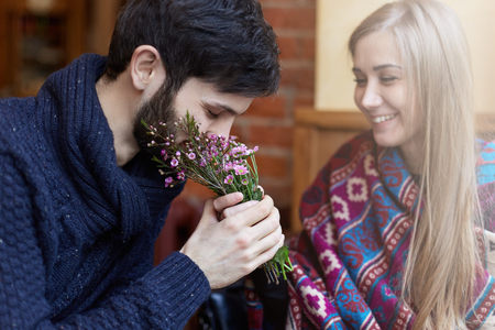 Focus on young handsome Caucasian man who is smelling fresh flowers on a date with young smiling beautiful lady. Wearing casual warm clothes the spend happy time together in a modern coffeshop.の写真素材