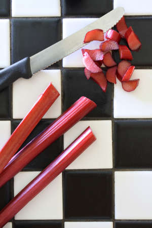 Three stalks of fresh garden rhubarb on a black and white tile counter, being cut with a serrated knife on a retro black and white tile counterの写真素材