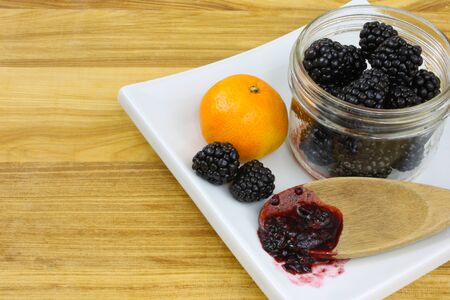 Blackberries in a jar, with a clementine orange and a wooden spoon of blackberry jam on a white plate sitting on a wooden countertop の写真素材