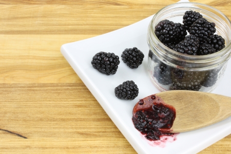 Blackberries in a jam jar, with three more blackerries and a wooden spoon holding blackberry jam, all on a white plate, set on a wooden counter top の写真素材