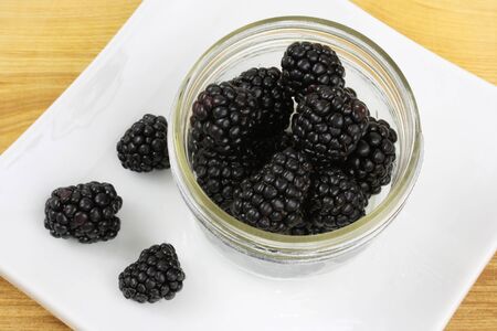 Blackberries in a jam jar, with three more blackerries, all on a white plate, set on a wooden counter top の写真素材