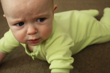 A baby in a light green outfit on a brown carpet, crying and looking angry の写真素材