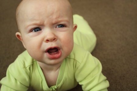 A baby in a light green outfit on a brown carpet, crying and looking angry の写真素材