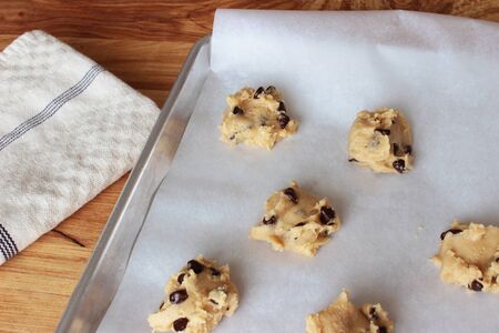 A close-up image of a cookie sheet lined with parchment paper, with chocolate chip cookie dough shaped into cookies, on a wooden counter with a white dish towel.の写真素材