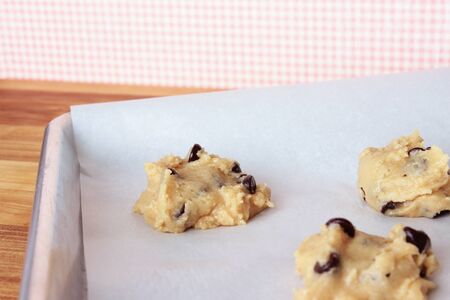 A close-up image of a cookie sheet lined with parchment paper, with chocolate chip cookie dough shaped into cookies, on a wooden counter with a pink and white gingham wallpaper background.の写真素材