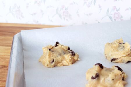 A close-up image of a cookie sheet lined with parchment paper, with chocolate chip cookie dough shaped into cookies, on a wooden counter with a vintage white wallpaper background with pink flowers.の写真素材