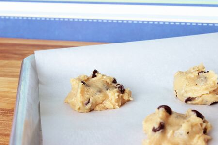 A close-up image of a cookie sheet lined with parchment paper, with chocolate chip cookie dough shaped into cookies, on a wooden counter with a modern blue striped wallpaper background.の写真素材