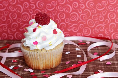 A lemon poppyseed cupcake with a swirl of buttercream icing and a raspberry on top, on a brown plaid tablecloth with heart shaped confetti and sprinkles, and pink and red ribbons, with a red background with pink swirls の写真素材