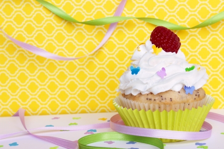 A lemon poppyseed cupcake with a swirl of buttercream icing, lemon zest, and a raspberry on top, on a yellow patterned background with butterfly confetti and sprinkles, and colorful ribbons   の写真素材