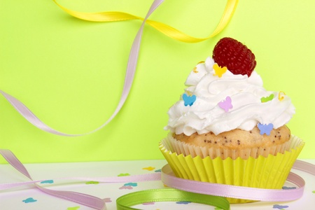 A lemon poppyseed cupcake with a swirl of buttercream icing, lemon zest, and a raspberry on top, on a green background with butterfly confetti and sprinkles, and colorful ribbons   の写真素材