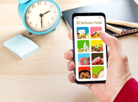 Male hand holding phone with app delivery food on screen above desk at home or officeの写真素材