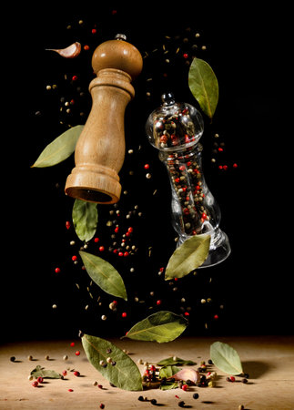 Two spice mills with peppercorns and laurel bay leaves on black flying over table.の写真素材