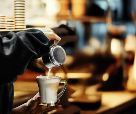 Barista pours rich, steamed milk into a glass cup, crafting stunning cappuccino art in a cozy cafe shopの写真素材