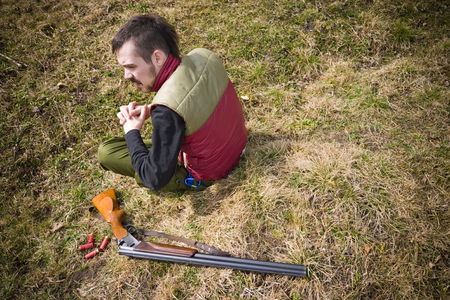 A view of a hunter sitting on the ground with a shotgun and several shotgun shells laying beside...
