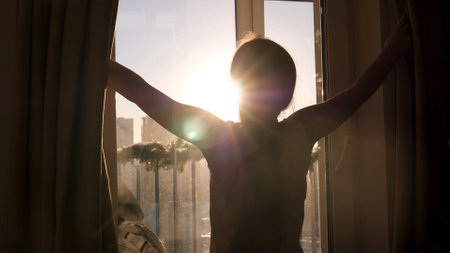 Young Girl Child Opens Curtains So Morning Sunlight Illuminates Cozy Apartmentの写真素材