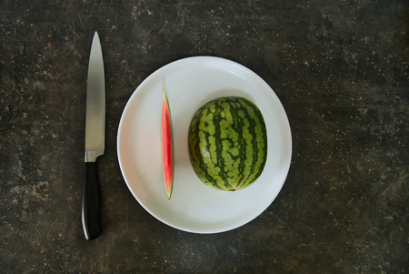 Half a watermelon on a white plate, next to a knife, both compositions make one ten eachの写真素材